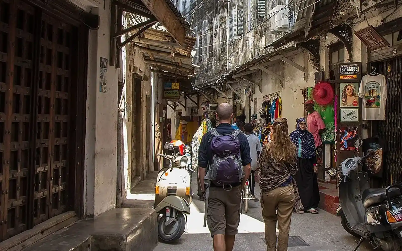 Reisende gehen durch eine enge Gasse in Stone Town, vorbei an Läden und einem Roller