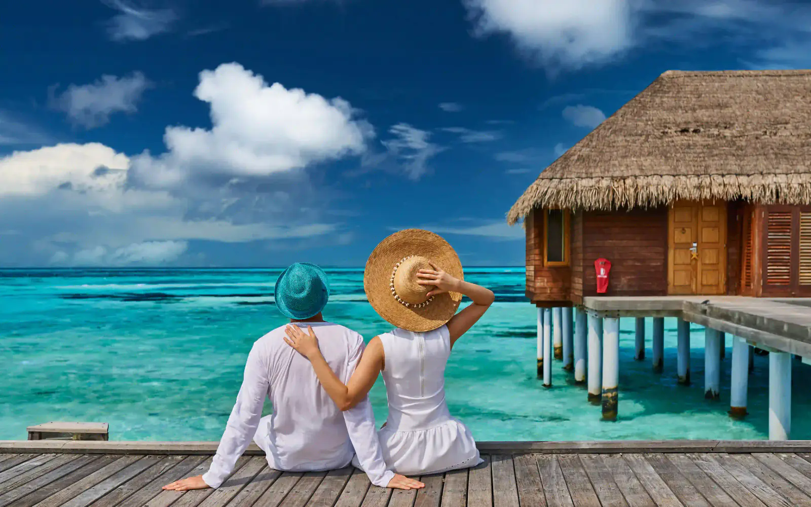 A couple in sun hats sitting on a wooden deck beside a thatched overwater cabin, turquoise sea ahead